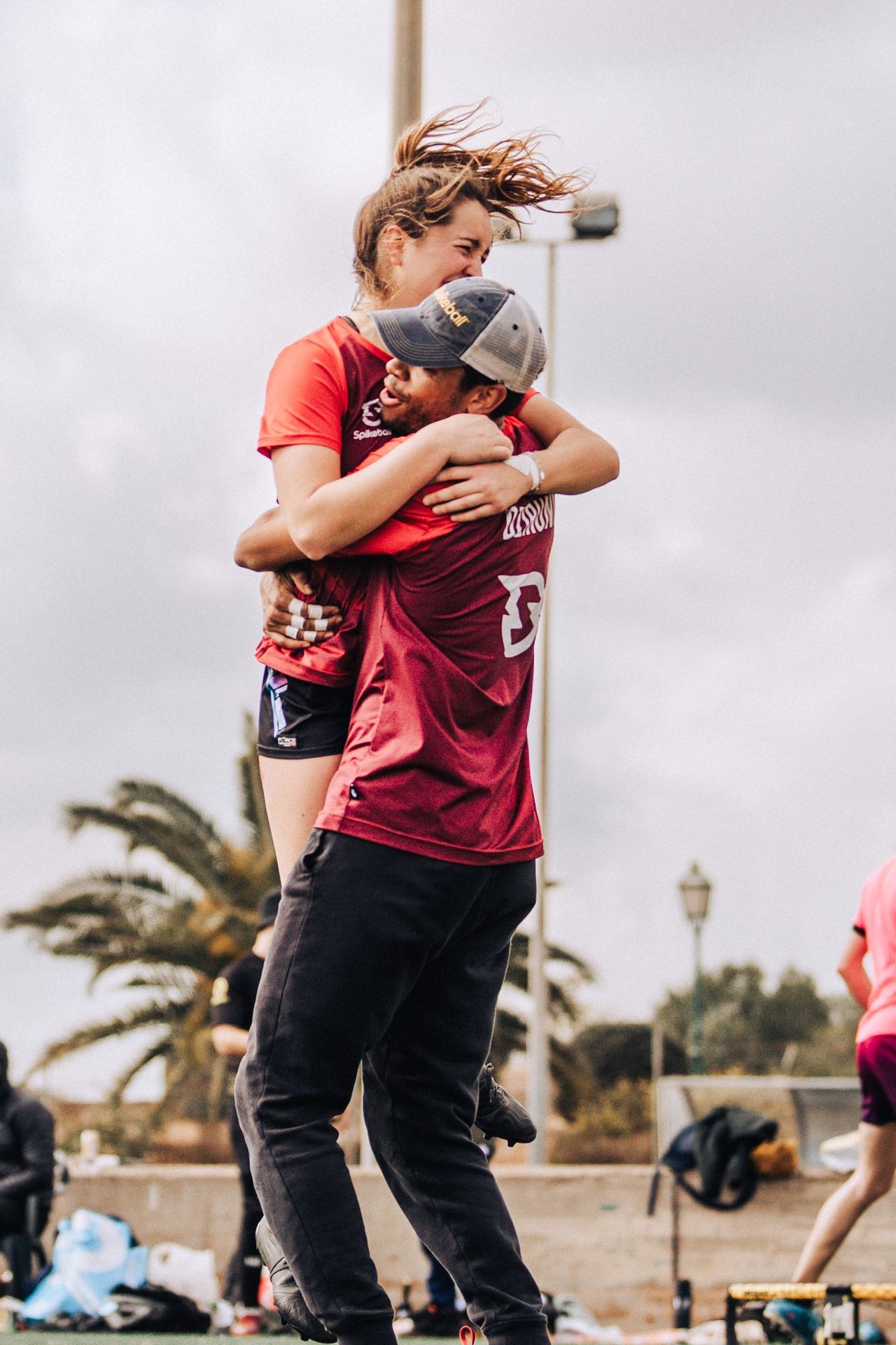 Two people wearing red sports jerseys embracing outdoors with palm trees and a cloudy sky in the background.