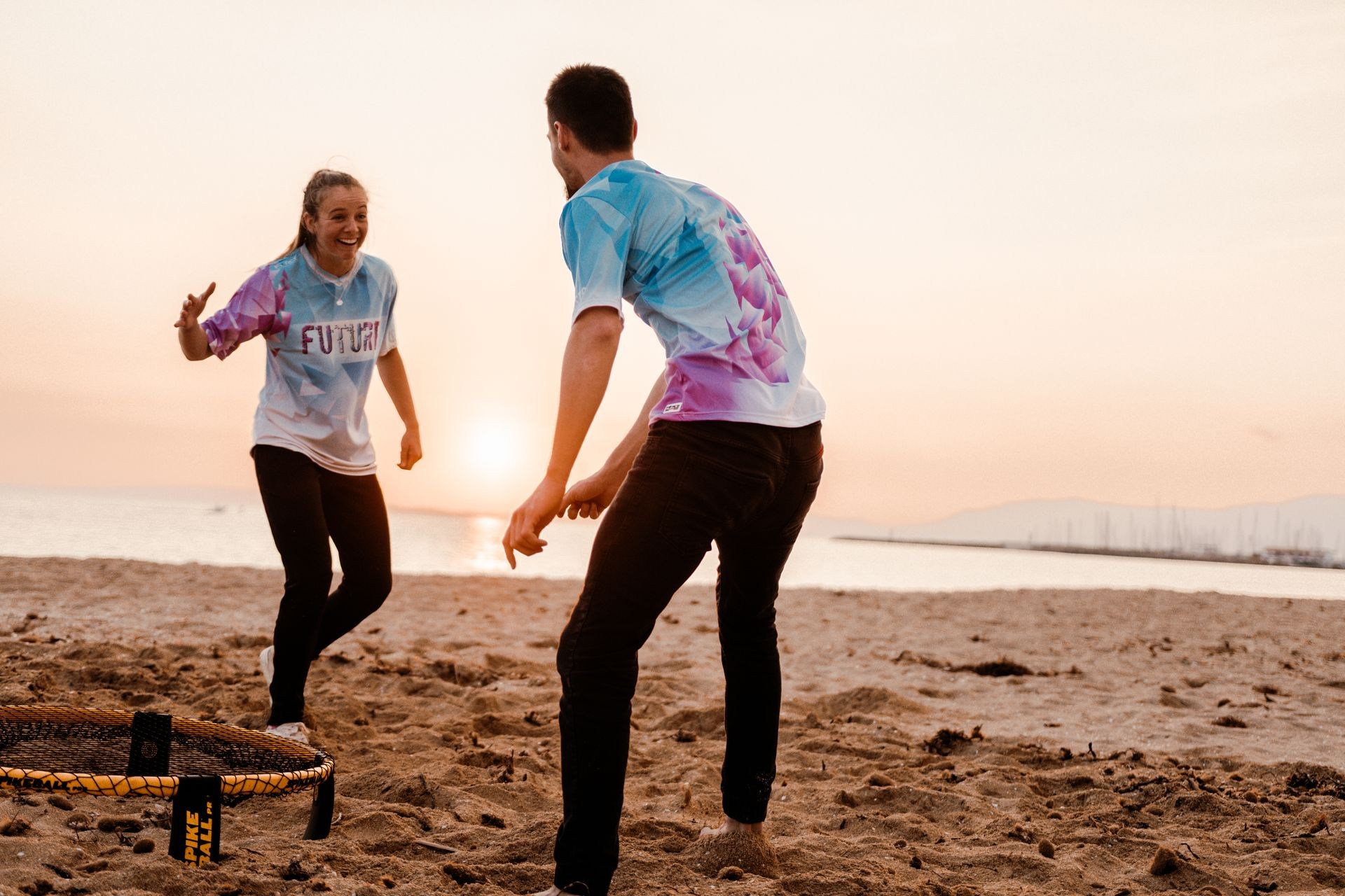 Two people playing Spikeball on a sandy beach at sunset, smiling and enjoying the game.